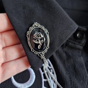 May include: A black and silver brooch featuring a black rose design. The brooch is oval-shaped with an ornate silver frame and a dangling chain. The brooch is attached to a black garment with white embroidery.