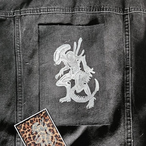 Pode incluir: Jaqueta jeans preta com um grande patch prateado com um gráfico alienígena nas costas. Uma patch retangular menor com estampa de leopardo e um gráfico alienígena também é visível.