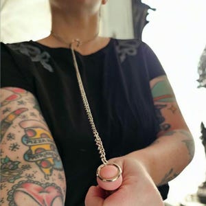 Op de afbeelding: Een zilverkleurige metalen ketting met een bijpassende ring aan het uiteinde. De ketting zit om de nek en de ring wordt in de hand gehouden. De persoon draagt een zwart shirt.