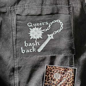 Pode incluir: Casaco de ganga cinzento escuro com um bolso com o texto branco "Queers bash back". O design inclui uma maça, uma corrente e uma espada. Uma pequena aplicação com estampa de leopardo com o mesmo design também é visível.