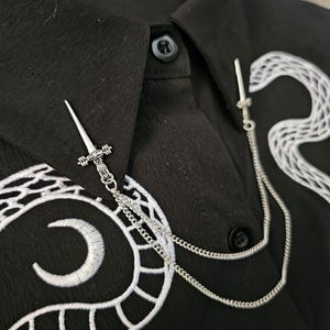 May include: A silver sword-shaped brooch with a chain, attached to a black shirt. The shirt has white embroidered detailing and a button-down collar. The brooch adds a decorative element to the garment.