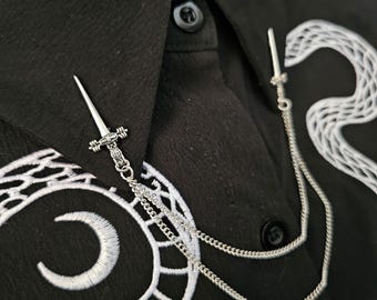 May include: A silver-colored brooch featuring a sword design, connected by a delicate chain. The brooch is attached to a black shirt with white embroidered detailing. The shirt has a button-down collar.