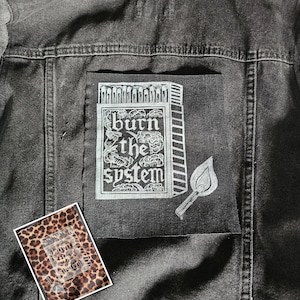 Pode incluir: Jaqueta jeans preta com um patch gráfico branco que diz "burn the system" com um design de caixa de fósforos e fósforo aceso. Um patch menor com estampa de leopardo com o mesmo design também é visível.