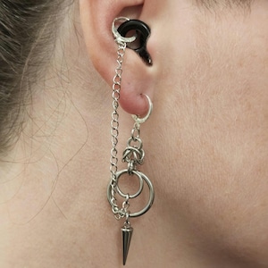 May include: A silver-toned ear jewelry set featuring a black circular cartilage ring connected by a delicate chain to a dangling earring. The earring includes a small hoop, a chainmail-style link, two silver rings, and a sharp, conical spike charm.
