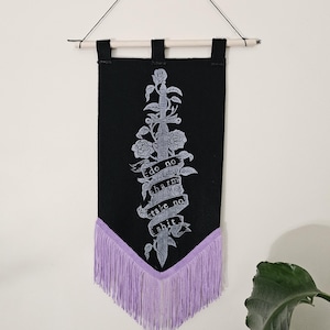 May include: Black wall hanging with a sword and rose design. The banner reads "do no harm take no shit" in white text. The bottom edge has lavender fringe, and it hangs from a wooden dowel.