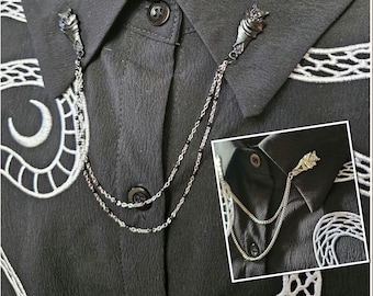 May include: A silver-toned collar chain with wolf head accents. The chain is attached to the collar of a black shirt with white embroidered details. The chain has two strands, one with black beads.