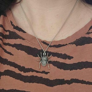 May include: Silver-toned spider pendant necklace. The spider charm has a detailed design with a textured body and legs. The necklace is worn on a person wearing a brown and black striped shirt.
