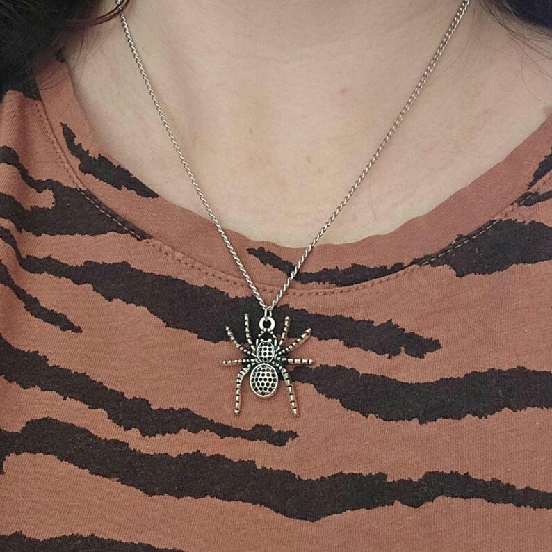 May include: Silver-toned spider pendant necklace. The spider charm has a detailed design with a textured body and legs. The necklace is worn on a person wearing a brown and black striped shirt.