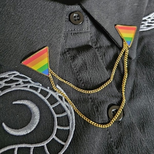 Op de afbeelding: Goudkleurige kraagspelden met een regenboogvlagontwerp. De driehoekige spelden zijn verbonden door een fijne gouden ketting. De spelden zijn bevestigd aan een zwart overhemd met decoratieve borduursels.