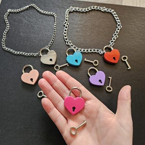 Può includere: Lucchetti a forma di cuore in vari colori, tra cui rosa, blu, rosso e viola, con chiavi abbinate. Alcuni lucchetti sono attaccati a collane a catena d'argento. Esposti su una superficie scura.