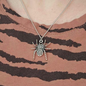 May include: Silver-toned spider pendant necklace. The spider charm has detailed legs and a textured body. The necklace is displayed on a person wearing a brown and black striped shirt. The chain is a delicate, silver-colored link.