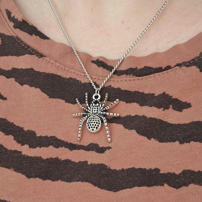 May include: Silver-toned spider pendant necklace. The spider charm has detailed legs and a textured body. The necklace is displayed on a person wearing a brown and black striped shirt. The chain is a delicate, silver-colored link.