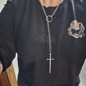 Puede incluir: Collar plateado con un colgante de espada. El collar tiene un diseño de cadena con un acento circular y un colgante de espada. La persona lleva una camiseta negra.