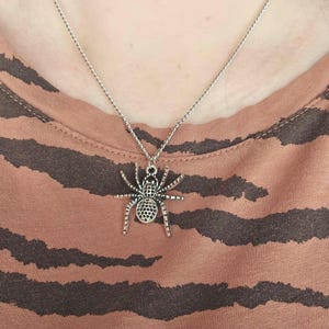May include: Silver-toned spider pendant necklace. The spider charm has detailed legs and a textured body. The necklace is worn on a person wearing a brown and black striped shirt.
