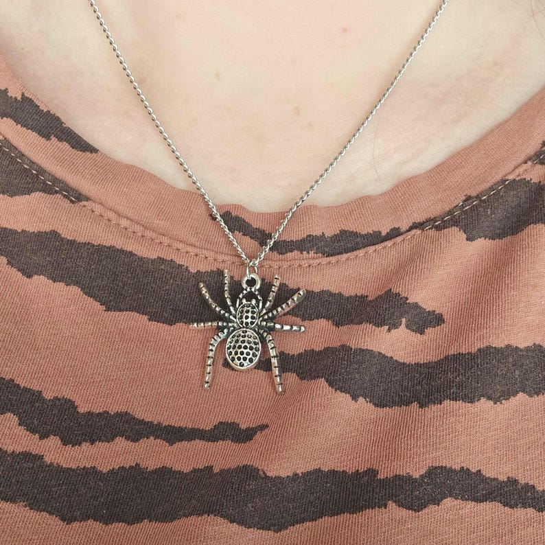 May include: Silver-toned spider pendant necklace. The spider charm has detailed legs and a textured body. The necklace is worn on a person wearing a brown and black striped shirt.