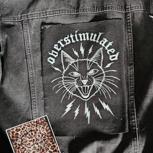 Pode incluir: Casaco de ganga preto com um grande patch com uma ilustração de gato branco e a palavra "oberstimulated". Um patch menor com o mesmo design em um fundo de estampa de leopardo também é visível.