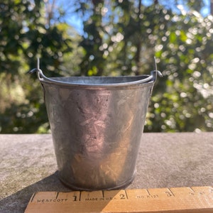 May include: A small, silver, metal bucket with a handle. The bucket is about 1 inch tall and is sitting on a wooden ruler.