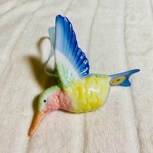 以下が含まれることがあります： 長いオレンジ色のくちばしとカラフルな体を持つセラミック製のハチドリの置物。翼は青と白、胸はピンク、体は黄色です。吊り下げ用の水色のリボンが付いています。置物の尾には小さな金のステッカーが付いています。