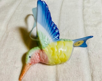 May include: A ceramic hummingbird figurine with a long, orange beak and a colorful body. The wings are blue and white, the chest is pink, and the body is yellow. A light blue ribbon is attached for hanging. The figurine has a small, gold sticker on its tail.
