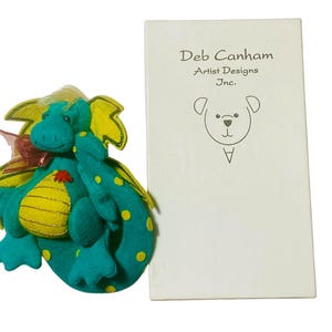 Peut inclure: Un dragon en feutre turquoise et jaune avec un nœud rouge et des pois jaunes. Le dragon est à côté d'une boîte blanche avec le texte "Deb Canham Artist Designs Inc." et une illustration d'ours.