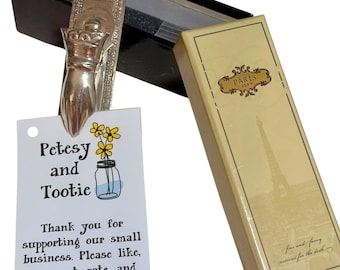 May include: A silver-toned metal clip shaped like a hand, attached to a white rectangular tag with the text Petesy and Tootie, a yellow flower illustration, and business information. Beside it is a cream-colored box with a Paris 1889 label.
