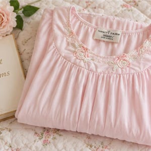 May include: A light pink nightgown featuring a round neckline with embroidered floral details in white and pink thread. The garment has a gathered bodice and a label inside the collar that reads Vanity Fair. The fabric is soft and opaque.
