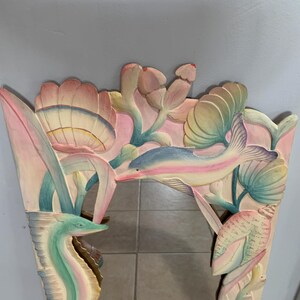 Op de afbeelding: Een gesneden houten spiegel met een fantasierijk onderwaterthema. De lijst heeft een dolfijn, zeepaardje en gestileerde bloemen in roze, blauwe en groene tinten.