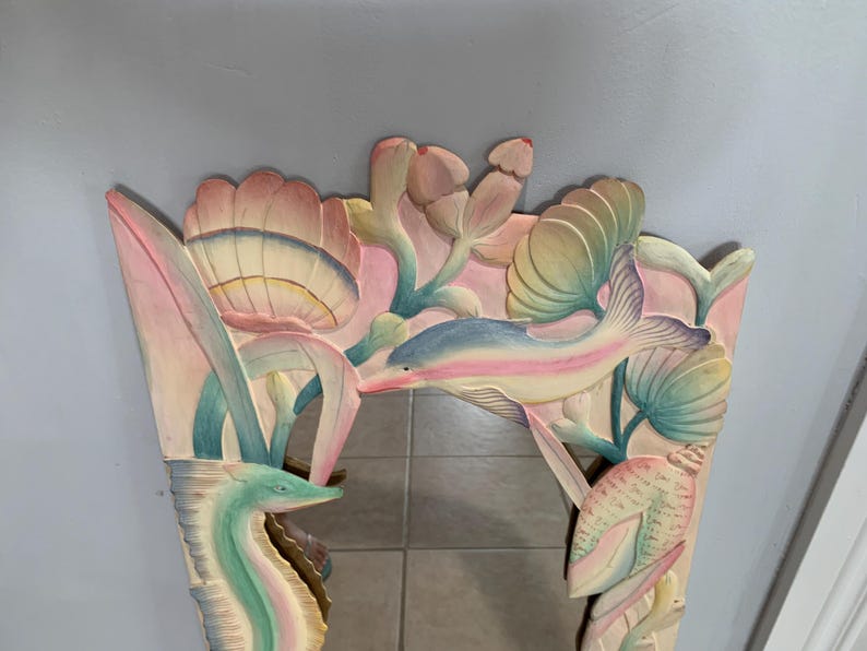 Op de afbeelding: Een gesneden houten spiegel met een fantasierijk onderwaterthema. De lijst heeft een dolfijn, zeepaardje en gestileerde bloemen in roze, blauwe en groene tinten.