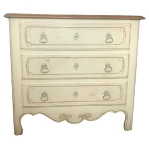 May include: A three-drawer chest featuring a cream-colored finish with distressed edges and a natural wood-toned top. Each drawer is adorned with two ornate metal ring pulls and a central decorative emblem, supported by curved cabriole legs.