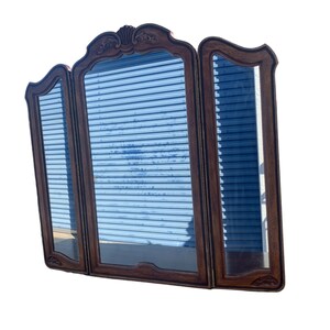 May include: A three-panel wooden mirror with a decorative carved frame. The mirror has a traditional style and is made of dark wood.