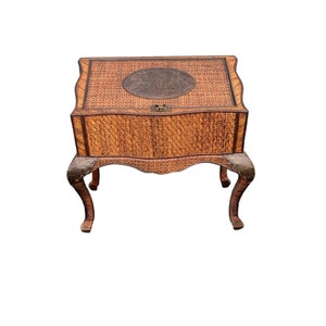 May include: A vintage, woven rattan side table with a square top and ornate, curved legs. The table features a dark brown, circular accent on the top and a latch closure. The woven texture and dark brown trim add to its classic appeal.