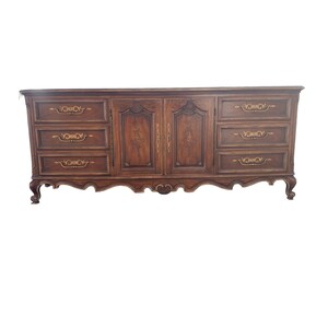 May include: A dark brown wooden dresser with six drawers and two doors. The drawers have ornate gold handles and the doors have a carved design. The dresser has a curved base and sits on four legs.