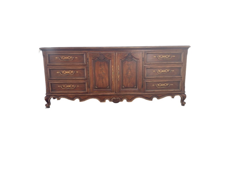 May include: A dark brown wooden dresser with six drawers and two doors. The drawers have ornate gold handles and the doors have a carved design. The dresser has a curved base and sits on four legs.