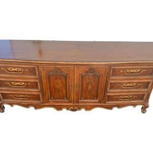 May include: A vintage wooden dresser with six drawers and two cabinet doors. The dresser has a curved front and ornate gold accents on the drawers and doors.