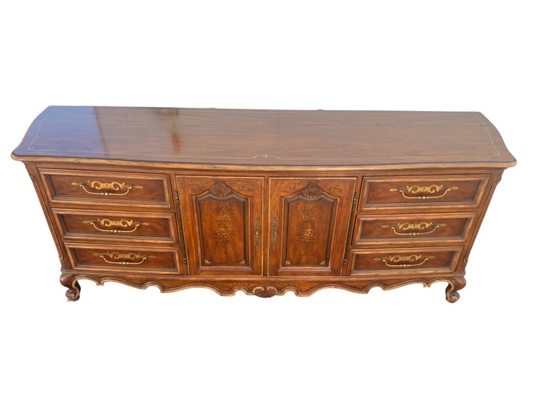 May include: A vintage wooden dresser with six drawers and two cabinet doors. The dresser has a curved front and ornate gold accents on the drawers and doors.