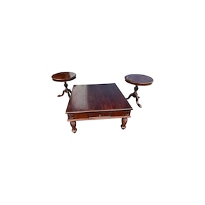 May include: A set of three dark brown wooden tables. The set includes a large rectangular coffee table with a drawer and two smaller round side tables. The tables have ornate, carved legs.