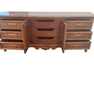 May include: A vintage wooden dresser with a dark brown finish and gold accents. It has six drawers with ornate handles and a glass top.