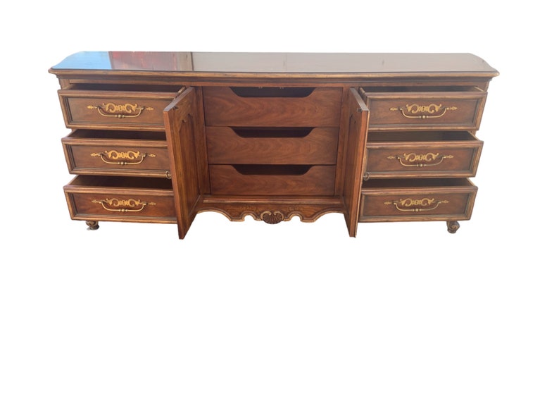 May include: A vintage wooden dresser with a dark brown finish and gold accents. It has six drawers with ornate handles and a glass top.
