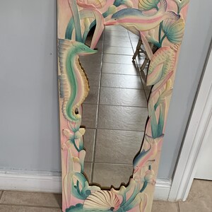 Op de afbeelding: Een gesneden houten spiegel met een ontwerp van een zeepaardje, schelpen en zeewier. De spiegel is geschilderd in tinten roze, blauw en groen.