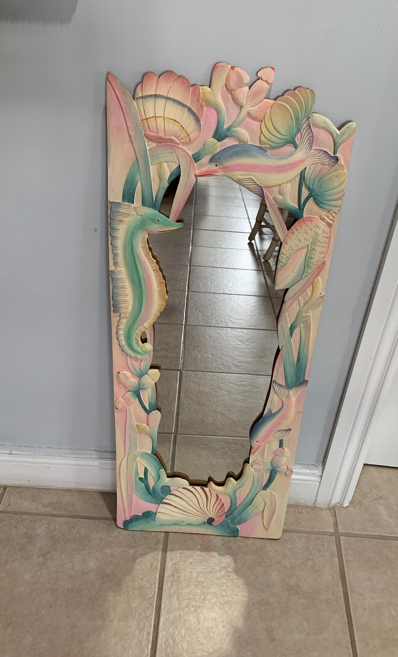 Op de afbeelding: Een gesneden houten spiegel met een ontwerp van een zeepaardje, schelpen en zeewier. De spiegel is geschilderd in tinten roze, blauw en groen.