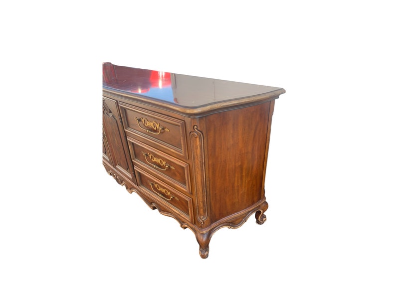 May include: A wooden dresser with six drawers and ornate gold-colored hardware. The dresser has a dark wood finish and a curved top.