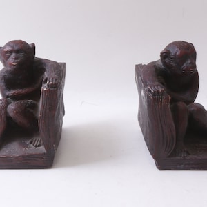 May include: A pair of dark brown wooden bookends shaped like monkeys sitting on books. The monkeys are holding the books with their arms and have a serious expression on their faces.