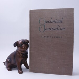 May include: A brown book with the title "Technical Journalism" by Peckman & O'Brien. The book is sitting on a white surface next to a brown pug figurine.