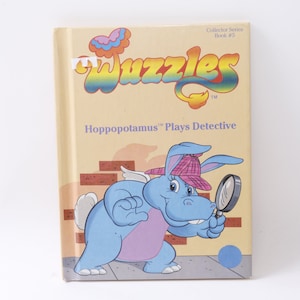Könnte beinhalten: Ein Buchcover mit einem blauen Flusspferd mit Flügeln und einem Detektivhut, das eine Lupe hält. Der Buchtitel lautet "The Wizzles" und der Untertitel lautet "Hippopotamus Plays Detective".