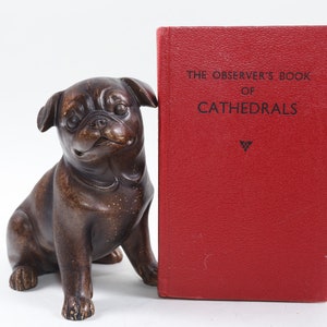 Könnte beinhalten: Ein rotes Buch mit dem Titel "The Observer's Book of Cathedrals" mit einem schwarzen Dreiecksymbol auf dem Einband. Das Buch lehnt an einer braunen Mopsfigur.