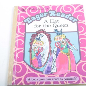 May include: A pink and blue book cover with a title that reads "Eager Reader A Hat for the Queen". The cover features an illustration of a queen with a cat on her head looking in a mirror.