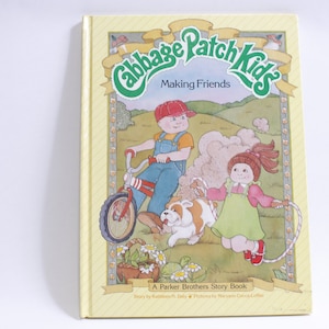 Op de afbeelding: Een gele boekomslag met een groene illustratie van twee Cabbage Patch Kids, een rijdt op een rode fiets en de ander springt touwtje. De titel van het boek is "Cabbage Patch Kids Making Friends" en de ondertitel is "A Parker Brothers Story Book".