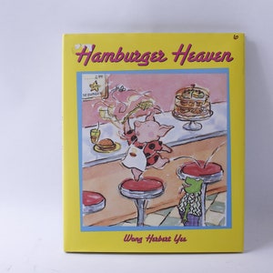 May include: A yellow book cover with the title "Hamburger Heaven" in a red and white font. The cover features a cartoon pig wearing a red and white polka dot dress jumping on a red stool. A green frog wearing a blue and white shirt is sitting on a red stool in the background. The book is by Wong Herbert Yee.