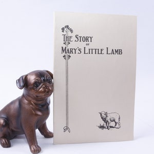 Könnte beinhalten: Ein Vintage-Buch mit dem Titel "The Story of Mary's Little Lamb" mit einer Schwarzweißillustration eines Lammes auf dem Cover. Das Buch liegt auf einer weißen Oberfläche neben einer bronzenen Mopsfigur.