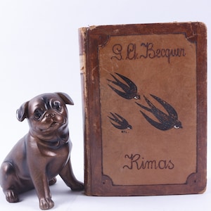 Puede incluir: Un libro encuadernado en cuero marrón con el título "Rimas" y el nombre del autor "G.L. Becquer" escrito en cursiva. La portada presenta una ilustración en blanco y negro de tres pájaros en vuelo.